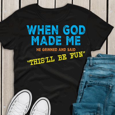 Black T-shirt with colorful text that reads, "When God Made Me, He Grinned and Said, This’ll Be Fun!" displayed flat with a bold, playful design.